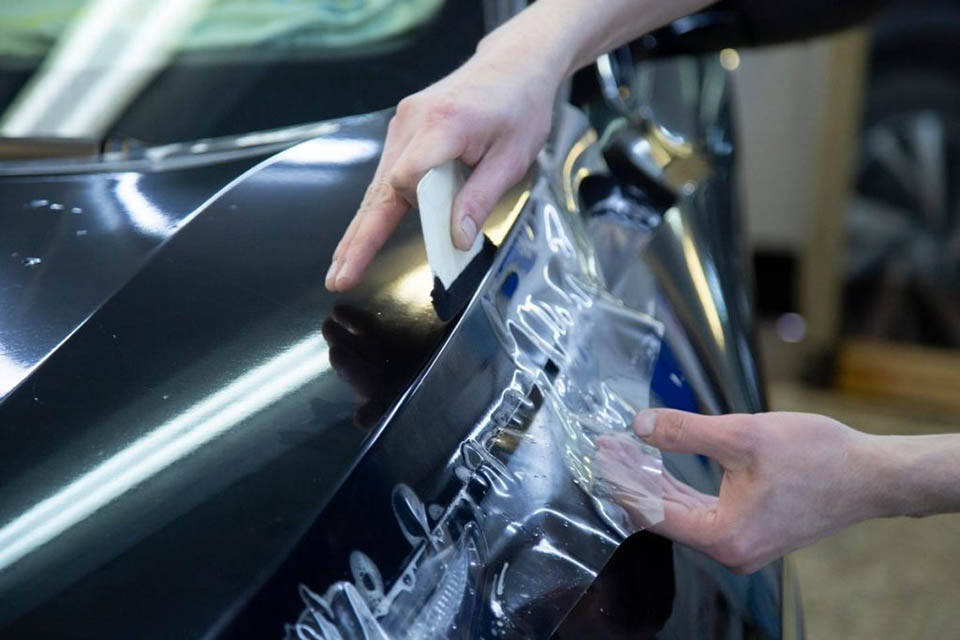 A technician using a squeegee tool to carefully apply transparent paint protection film onto the dark hood of a vehicle.