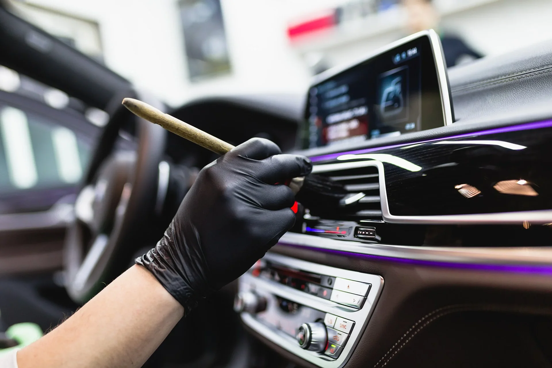 A close-up view of a technician wearing black gloves using a detailing brush with a wooden handle to clean the air vents and dashboard of a luxury vehicle interior, paired with interior surface protection.