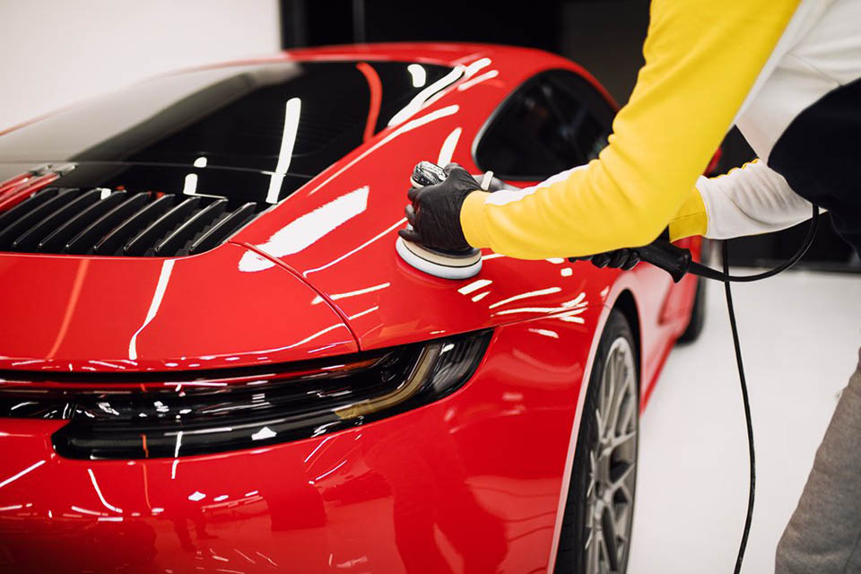 A professional detailer using a polishing machine on the rear body panel of a glossy red sports car in a clean garage.