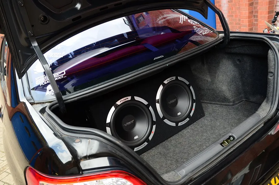 Dual 12-inch subwoofers in a carpeted enclosure box installed in the trunk of a dark car equipped with rear window tinting and decals.