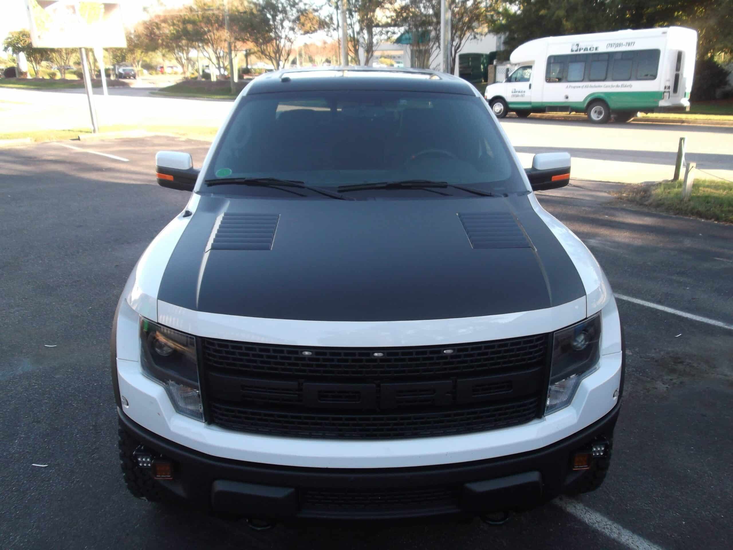Front view of a white Ford truck featuring a custom black hood vinyl wrap and window tint parked in a lot during daylight.