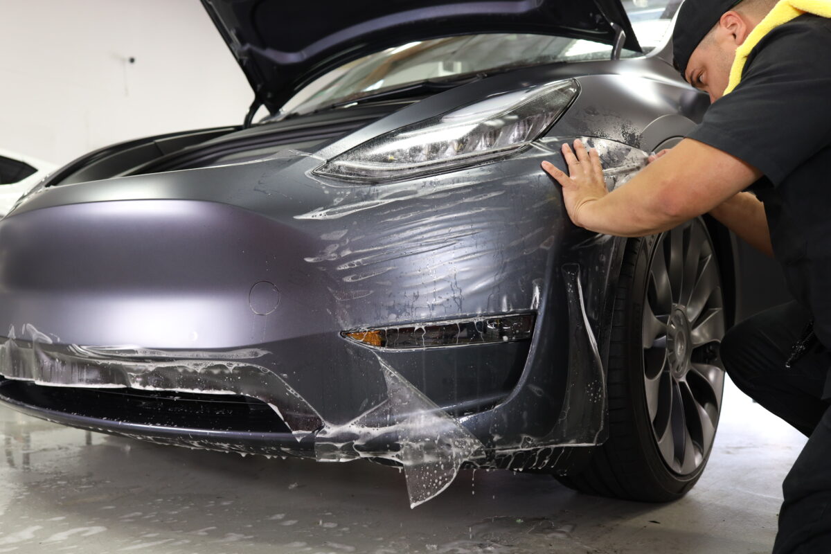 A professional technician applying clear paint protection film (PPF) and custom wrap to the front bumper of a sleek dark vehicle in a well-lit workshop.